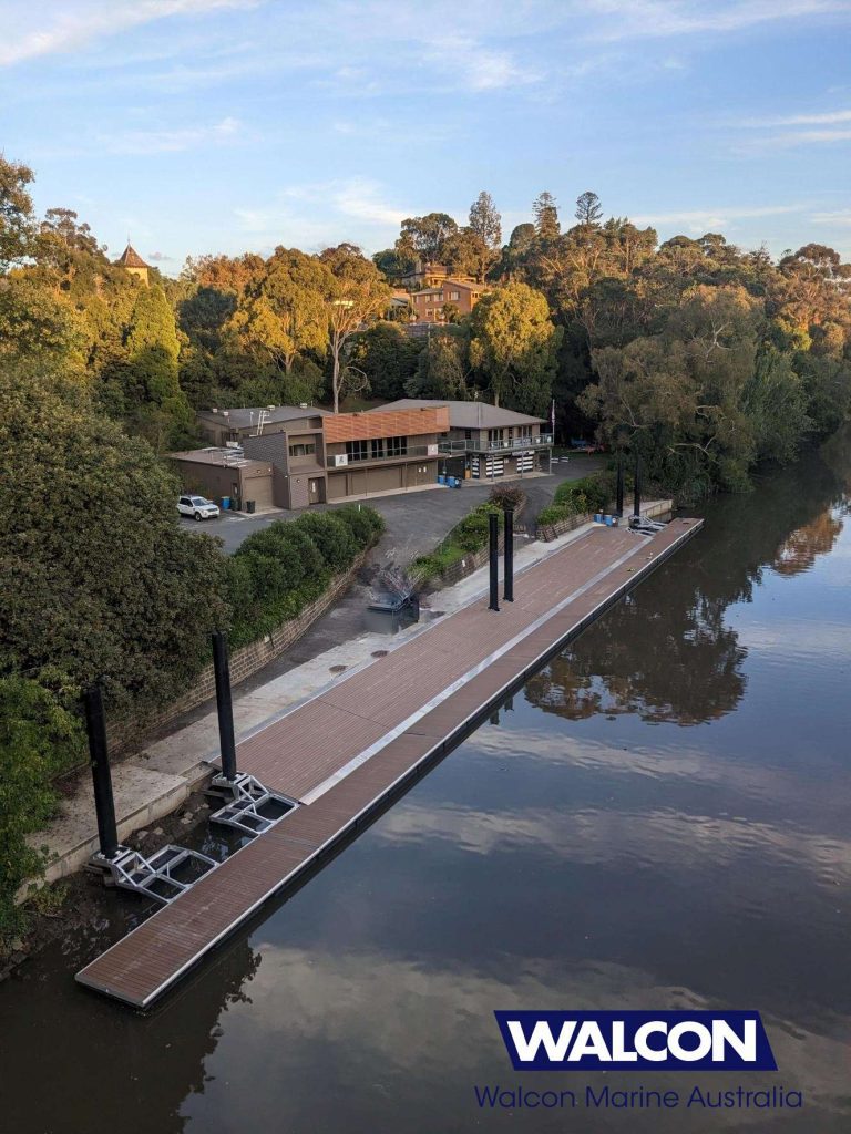 A serene river scene features a wooden dock stretching alongside the water, complemented by sturdy concrete pontoons. Surrounded by lush trees, a building stands nearby under a clear sky. The logo "WALCON Marine Australia" is visible in the bottom corner.