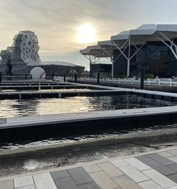 Modern buildings by a reflective water feature, with floating pontoons gently drifting under a setting sun.