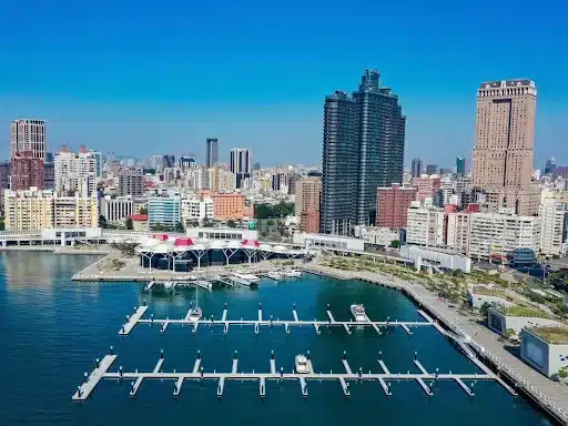 Aerial view of a city skyline with a marina in the foreground, showcasing aluminium pontoons and floating breakwater. Several boats are docked at the piers. Tall buildings and skyscrapers are visible, set against a clear blue sky.