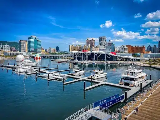 A marina filled with boats and yachts, set against a cityscape backdrop of modern buildings. Aluminium marinas gleam in the bright blue sky, where scattered clouds float above. Walkways and piers extend into the water alongside an innovative floating breakwater, ensuring calm waters.