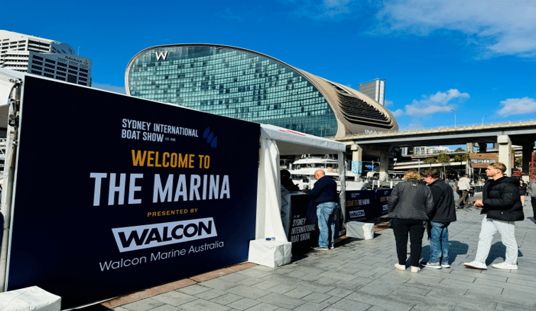 Outdoor event with a "Welcome to The Marina" sign at the Sydney International Boat Show, showcasing the craftsmanship of leading marina builders. People stroll on the pavement near a modern building with a unique curved design under a clear blue sky.