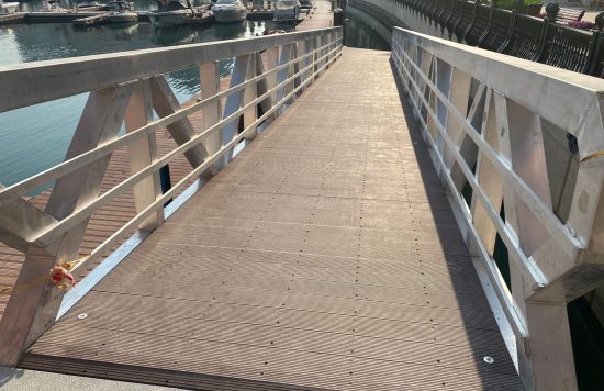 A sunlit wooden ramp leads to a dock, where boats are moored against sturdy concrete pontoons.